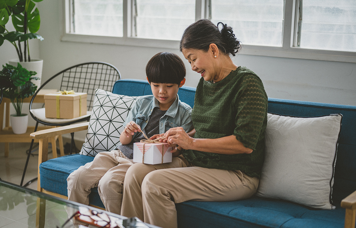 A grandmother gives a gift to their grandchild.  They are sitting together and the grandchild opening a gift.