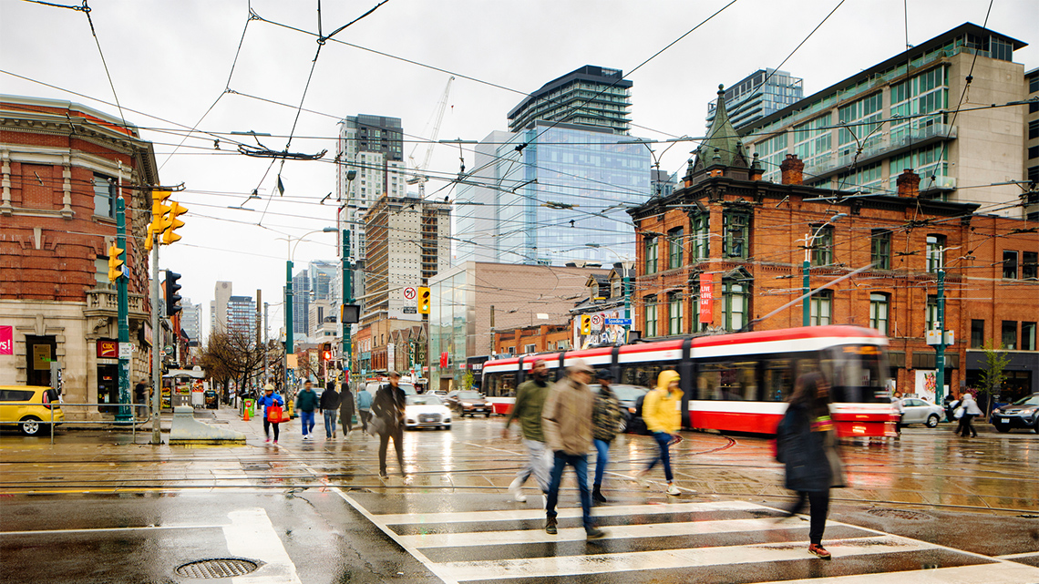 People at a crosswalk waiting to cross the a busy street with cars and a street car.