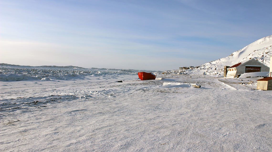 Iqaluit Beach in Nunavut in the winter. On the right there is a small snow hill with a small outhouse. In front of the house there is a small boat on the ground. The ground is covered in snow.