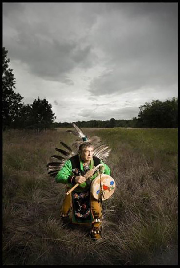 Indigenous elder wears traditional regalia and holds an instrument while storytelling.