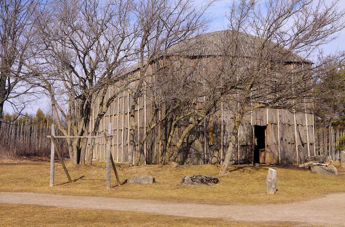 The image shows a photo of a Haudenosaunee longhouse with a rounded roof and with wood framing.