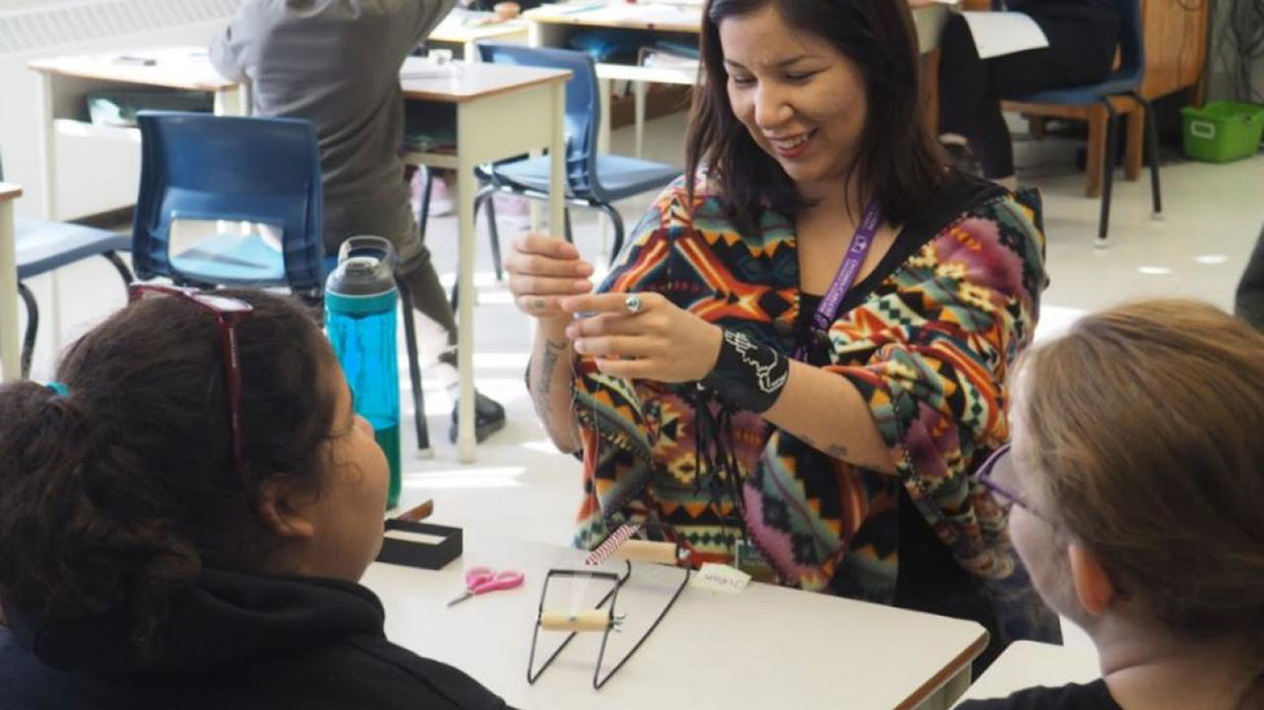 An adult leading a beading workshop in a classroom. There are two students in front of the adult sitting at a table.