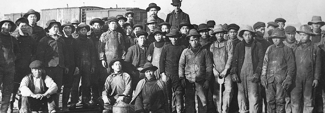 About 30 Chinese men working in BC on the railroad. They are wearing dusty overalls, long sleeve shirts, and hats.