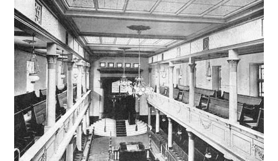 The inside of a synagogue. The ceiling is high with small chandeliers hanging on top. There is seating on the top and bottom level.