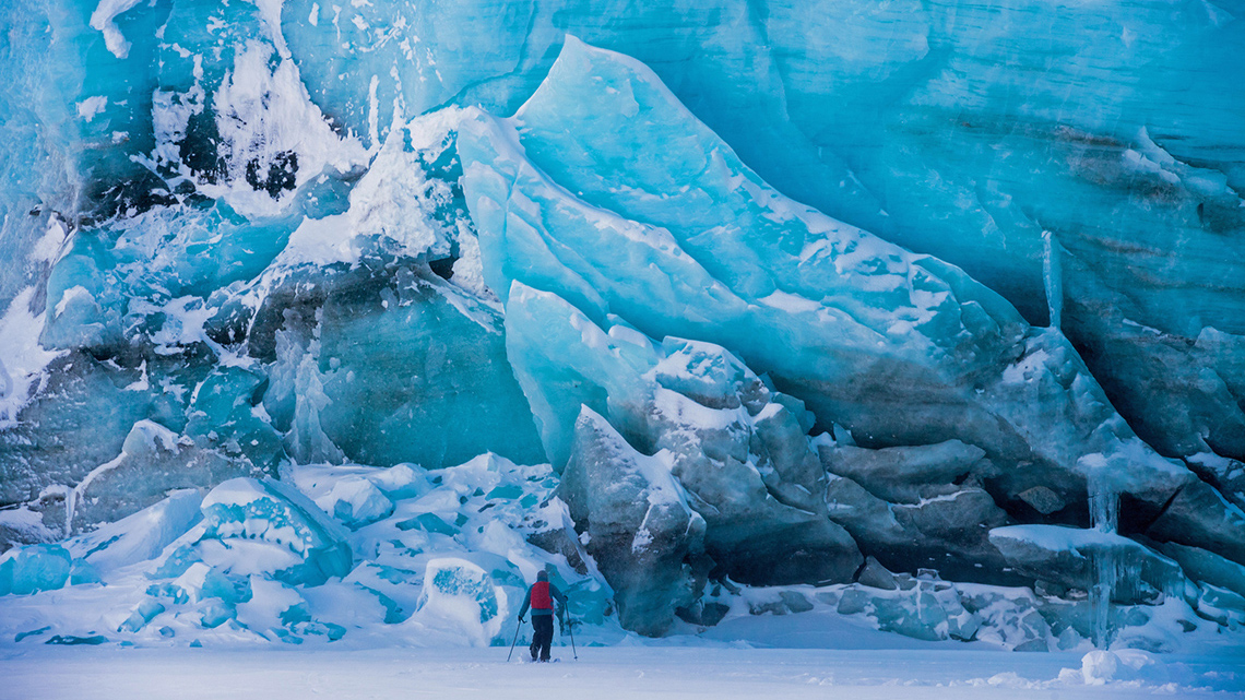 A person wearing a backpack skies past a huge glacier.