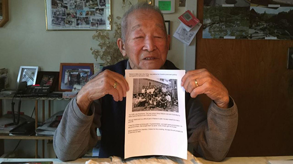 A photo of an elderly Japanese Canadian man holding a photo of a road construction camp.