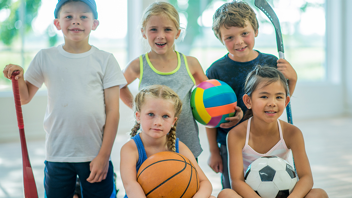Kids holding various types of sports equipment. They are inside a gym.