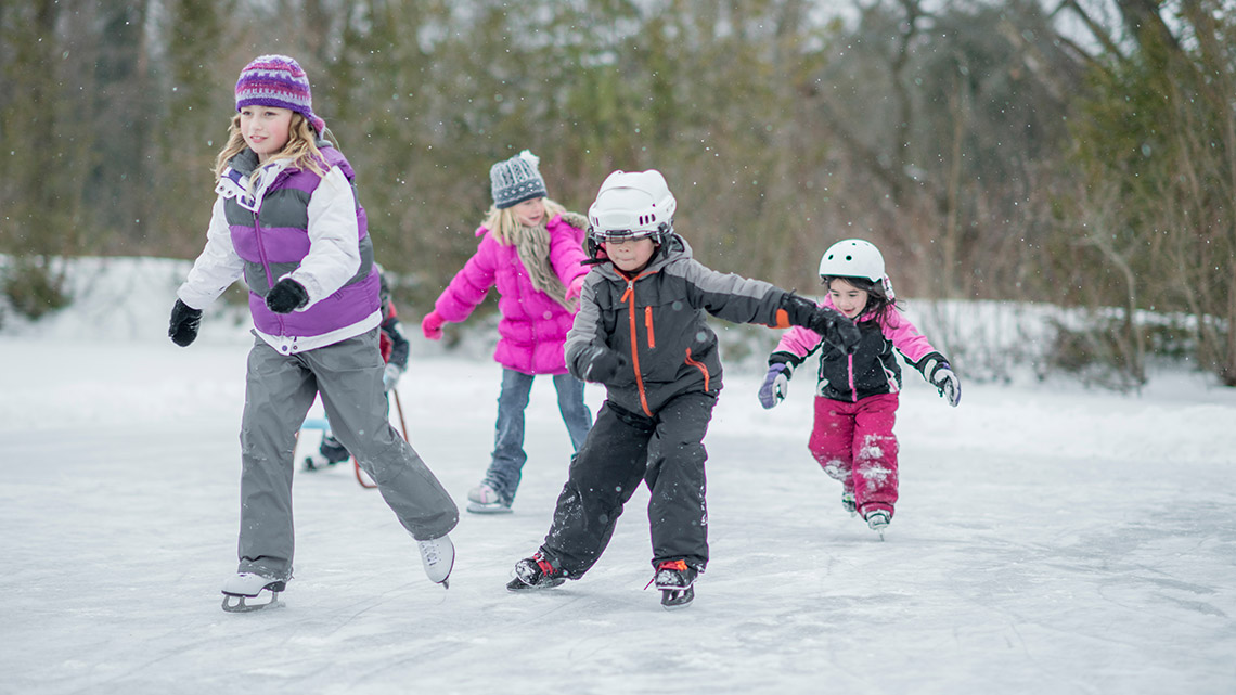 Four school age kids dressed in winter clothing, skating on an outdoor frozen pond.