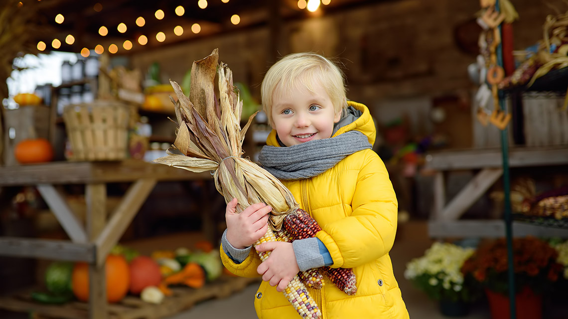 A local farmers' market. A child holding corn. There are stalls selling fruits and vegetables.