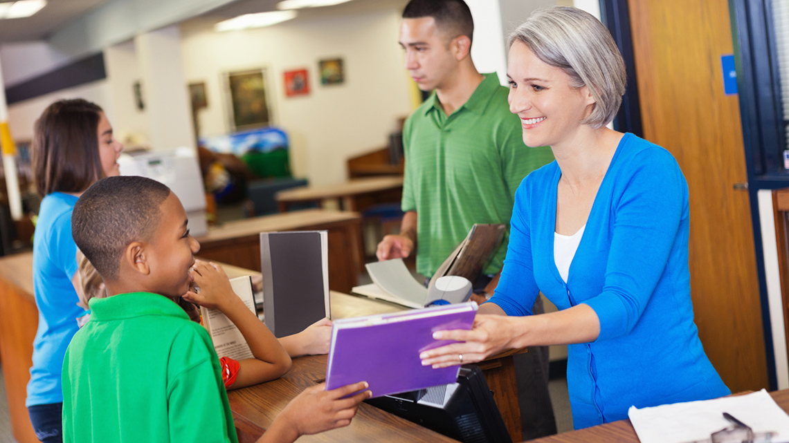 A young adult borrowing a book from the librarian, who is passing the book to them.