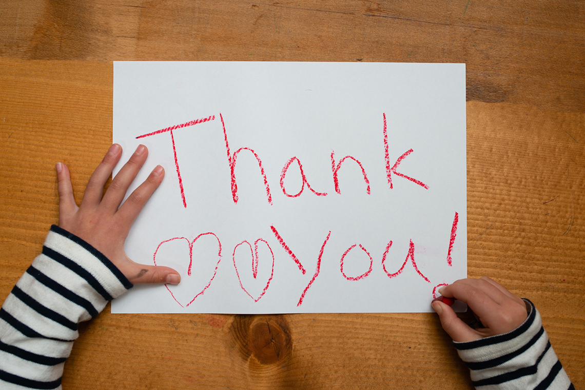 A set of hands writing the words “Thank you” on a piece of paper with a crayon.
