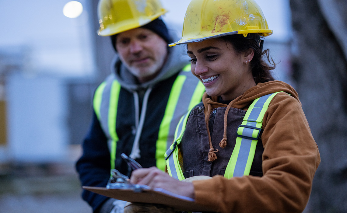 Two construction workers on a site. They are both looking at a clip board.