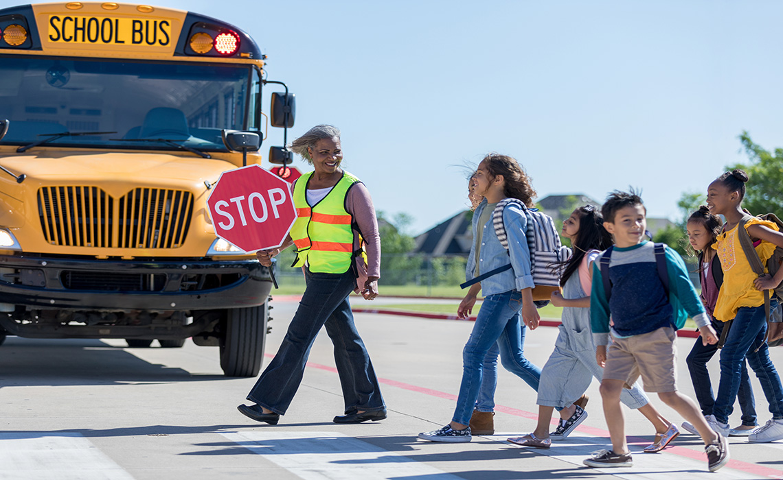 A crossing guard is standing in front of a school bus holding a stop sign as school age children cross the road.