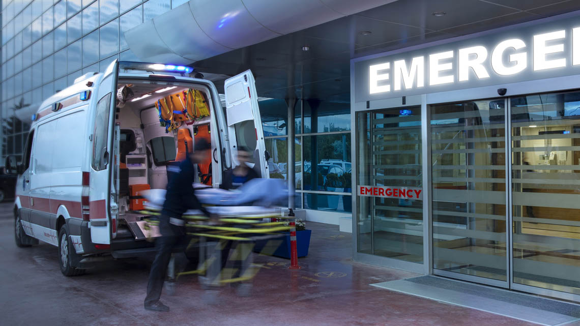 The front of the emergency department at a hospital. There is an ambulance out front with two paramedics pulling out a stretcher.