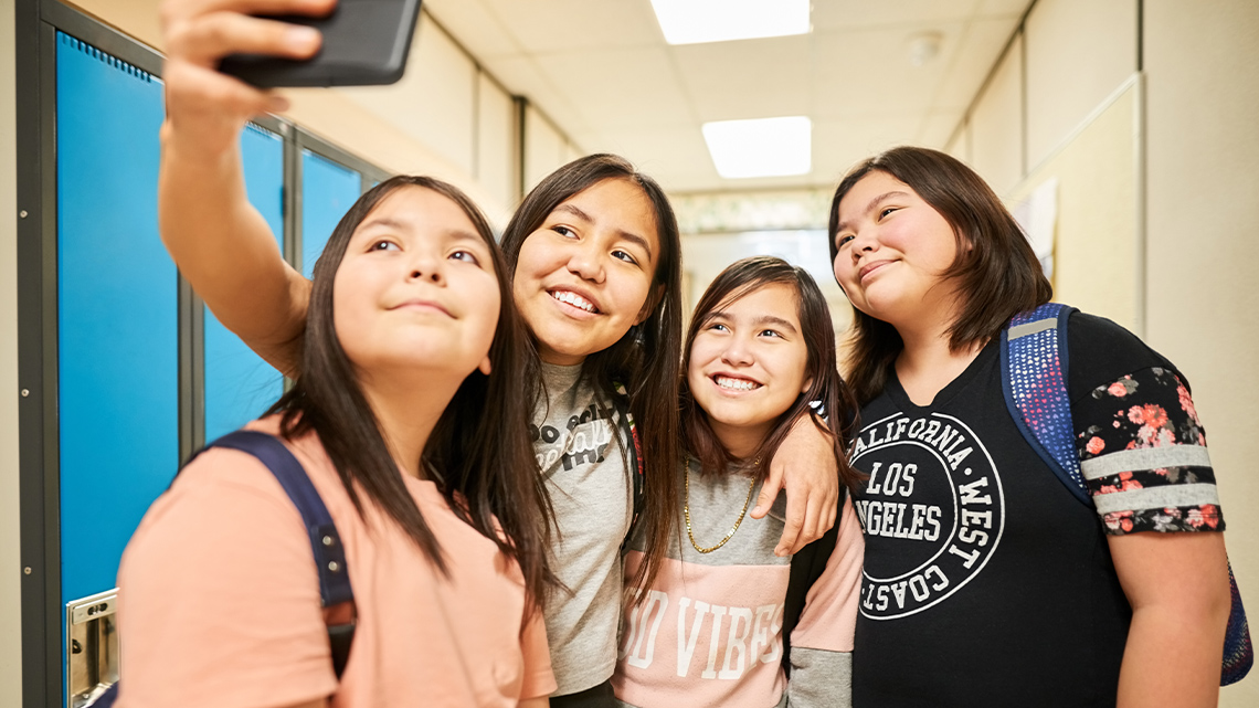 Four preteen students in a hallway. They are all looking at a phone taking a selfie.