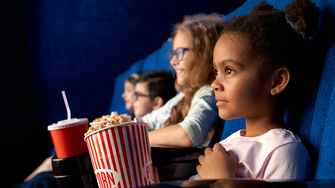 Four school age children sitting in a movie theatre staring at the big screen. They all have popcorn and sodas.