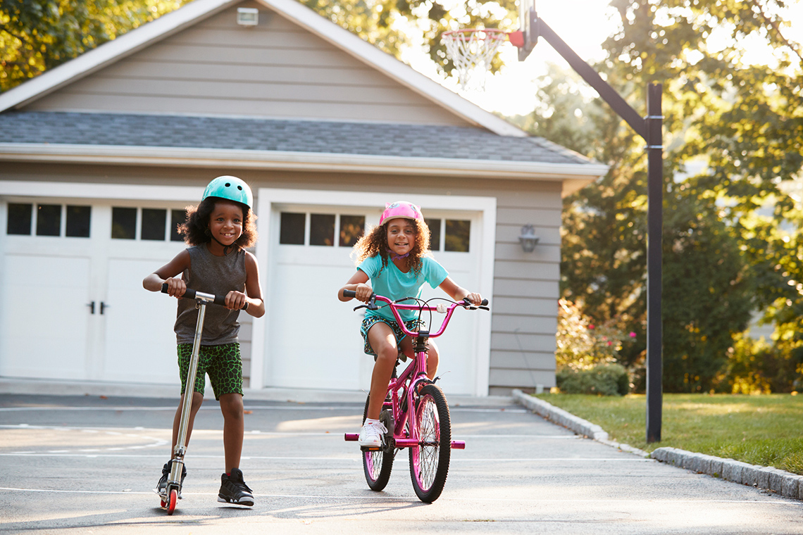 Two young children wearing helmets on a driveway. One child is on a scooter and the other is on a bike.