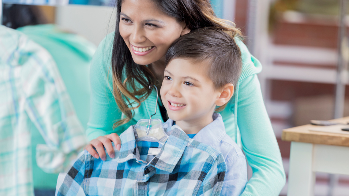 A child and a parent shopping at a clothing store. The parent has a shirt in front of the child.