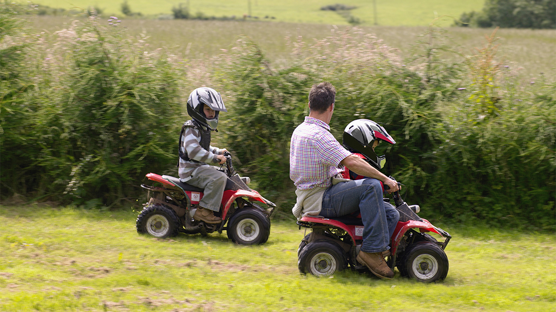 A child is driving an ATV. There is also another child driving an ATV with an adult.