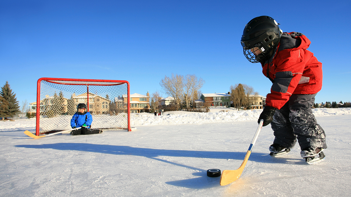 Two kids playing ice hockey on and outside rink.