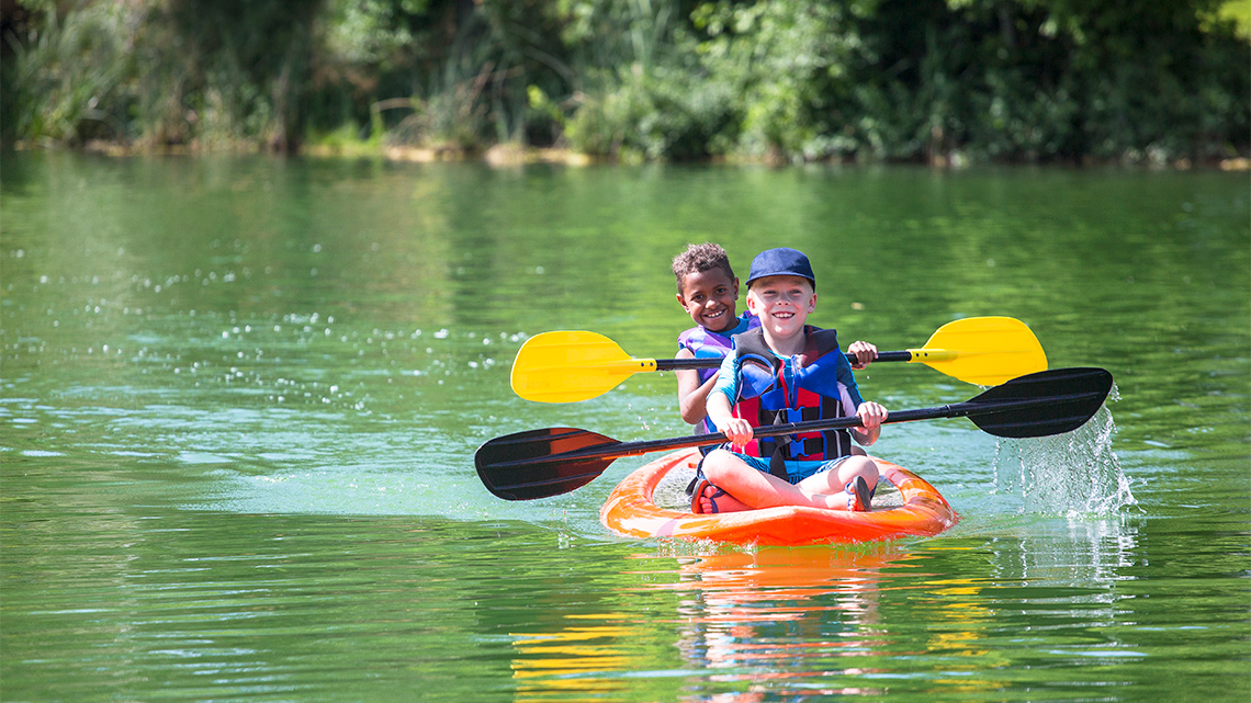 Two kids wearing life jackets are kayaking in a lake. There are trees in the background.