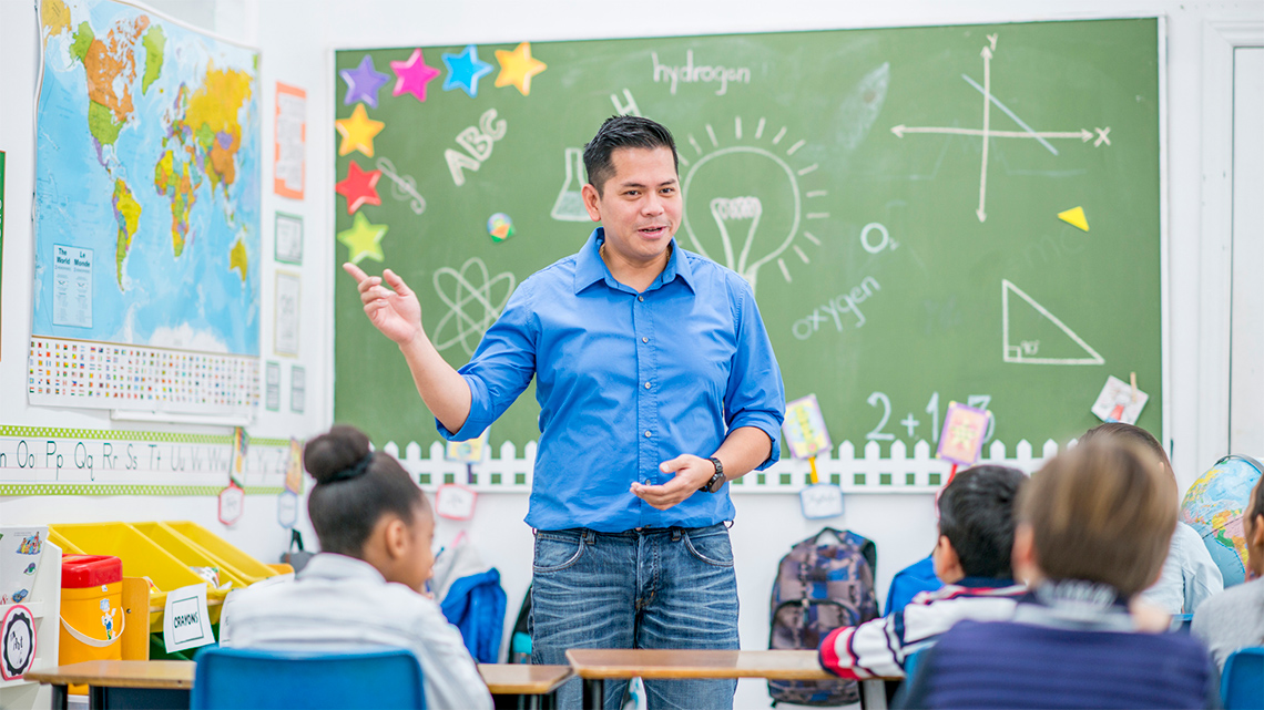 A teacher facing students in the classroom. There is a large chalkboard behind them.