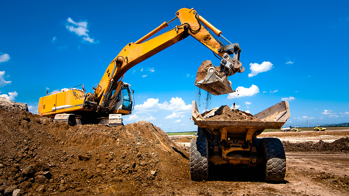 An excavator dumps dirt into the back of a large truck.