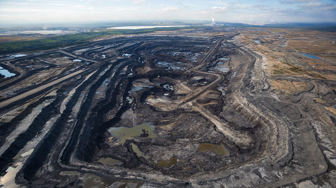Aerial view of a large open pit mine and small bodies of wastewater with a refinery in the background.