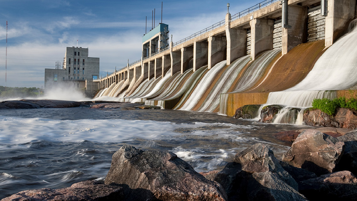 Water is being released through a dam into a large river with a power plant in the background.