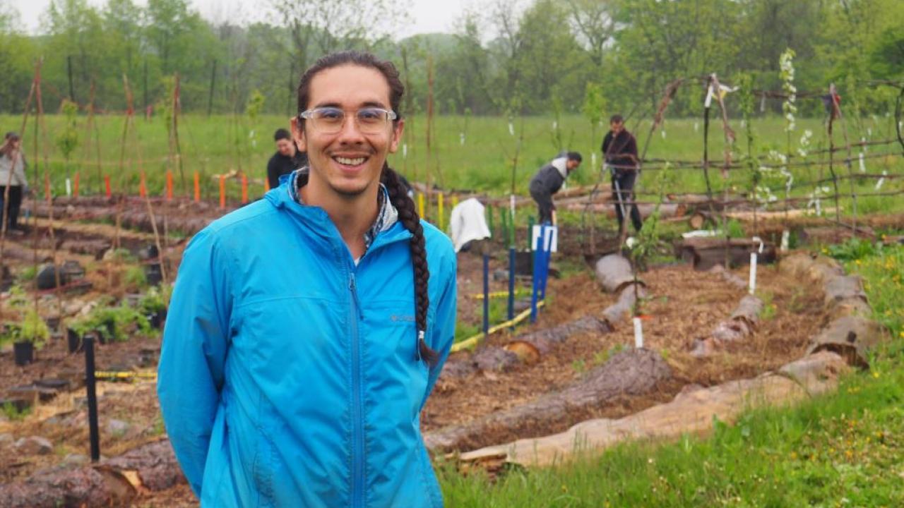 An adult smiling at the camera. There are people in the background working in a garden.