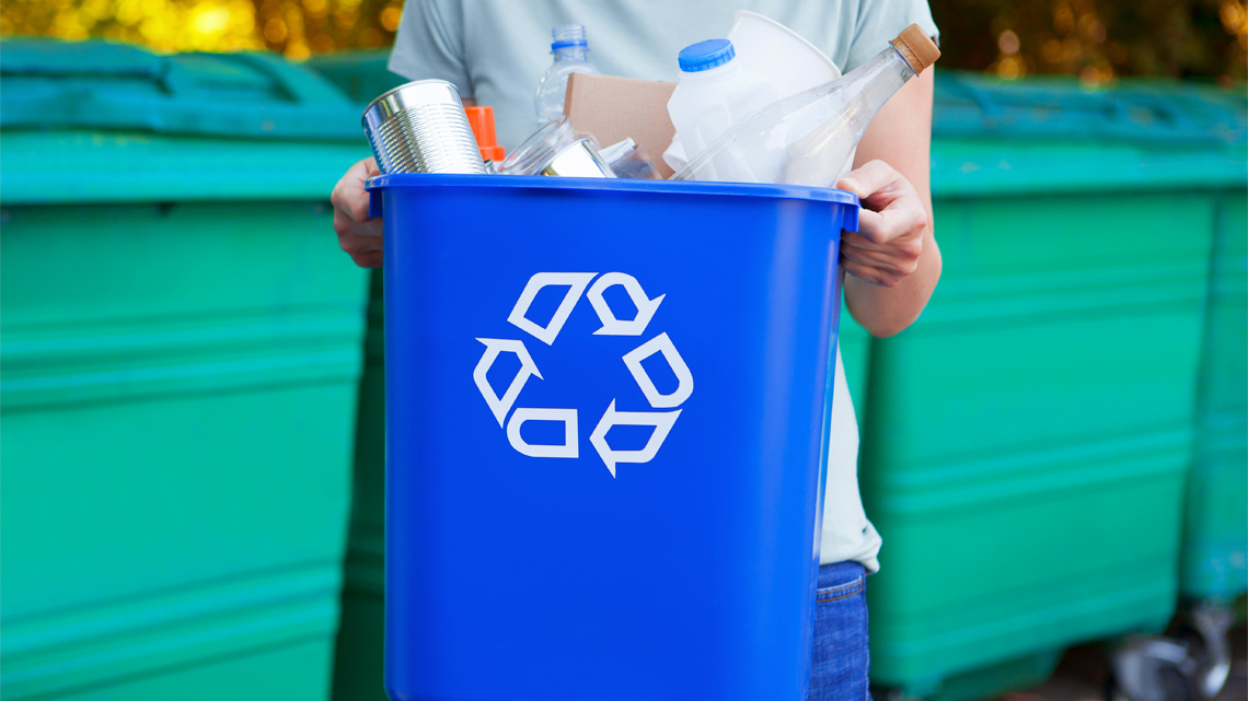 A person holding a blue recycling bin filled with products that can be recycled.